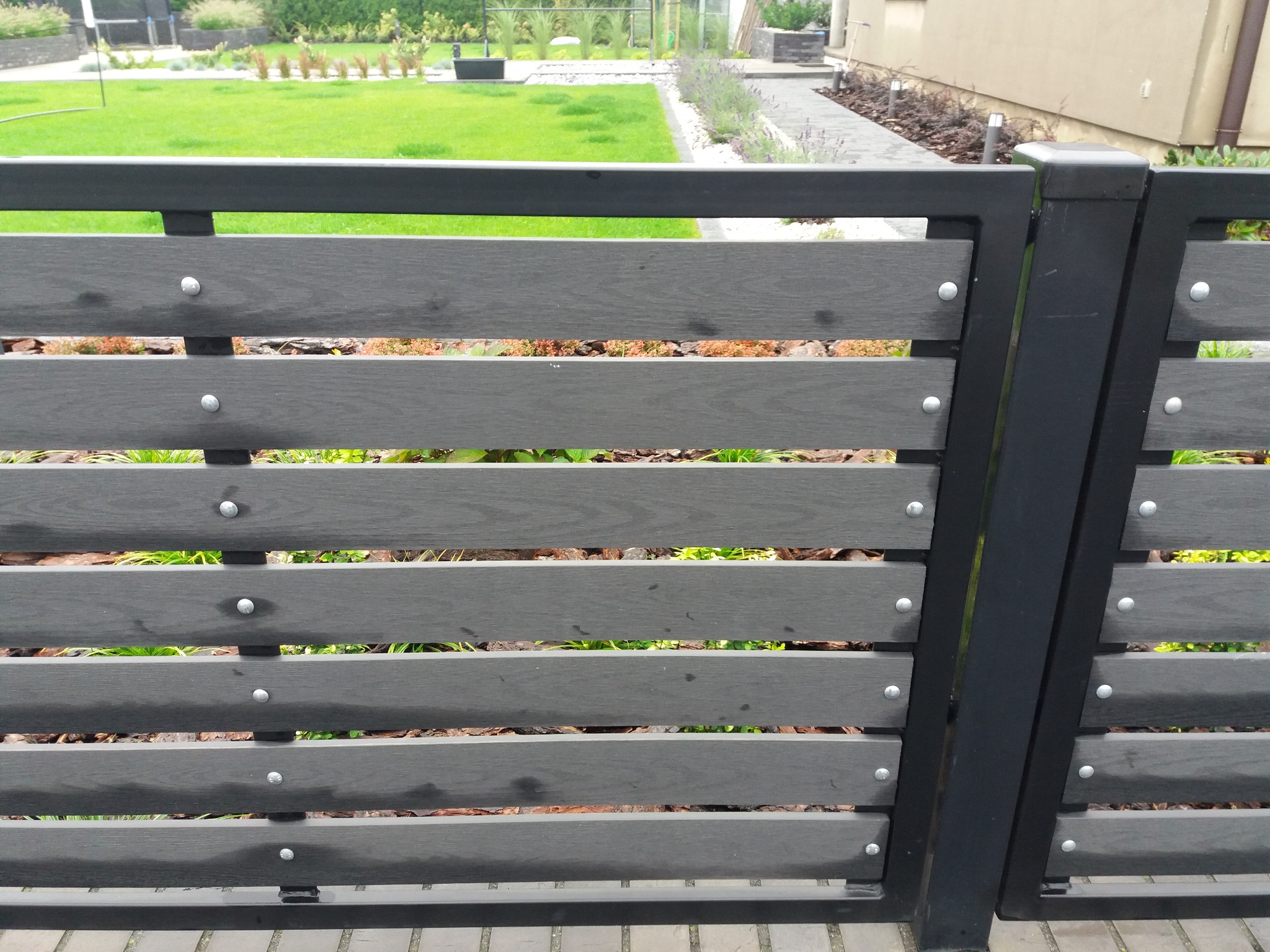 Nowoczesne ogrodzenie z paneli kompozytowych w odcieniach szarości, zamocowane na czarnych słupkach, z widocznymi srebrnymi nitami, na tle zadbanego trawnika i roślinności.