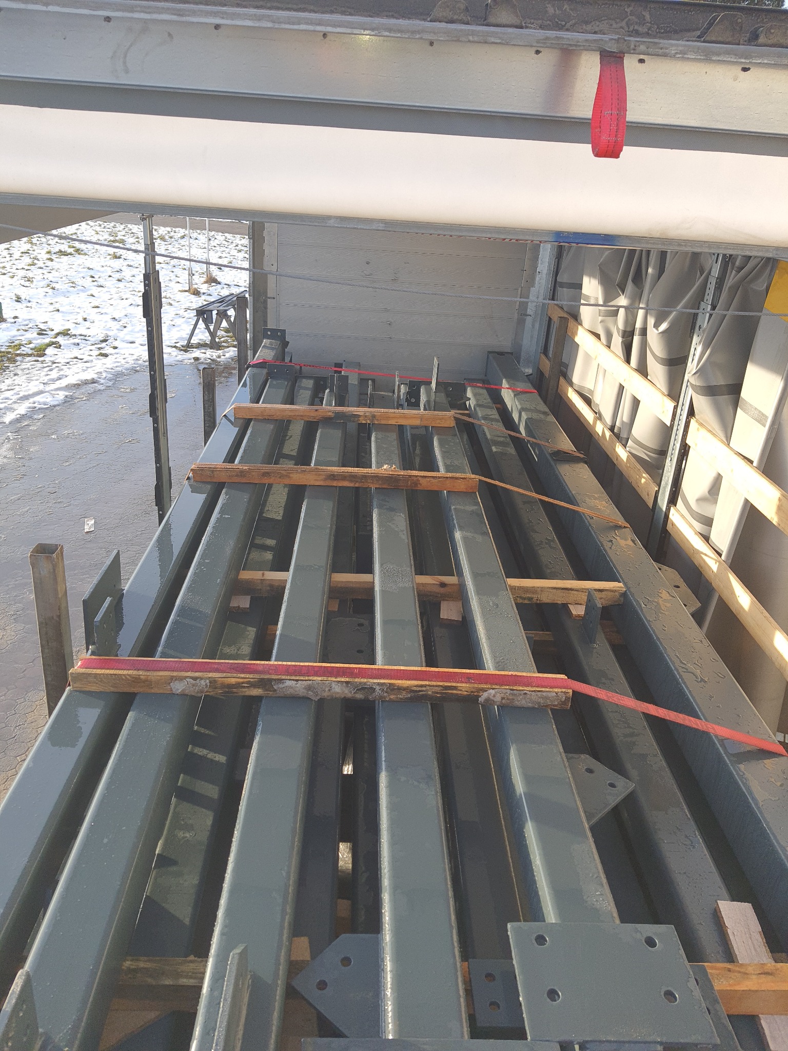 Szare, metalowe profile konstrukcyjne, zabezpieczone drewnianymi przekładkami i czerwonymi pasami transportowymi, wewnątrz naczepy ciężarówki.