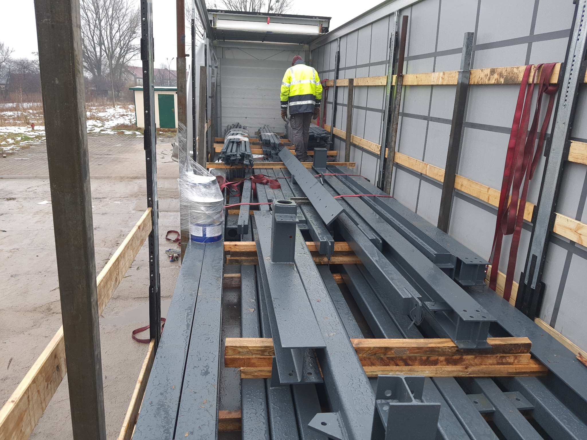 Załadunek pomalowanych na szaro stalowych belek konstrukcyjnych wewnątrz naczepy ciężarówki, zabezpieczonych drewnianymi przekładkami i pasami transportowymi, widoczny pracownik w odblaskowej...