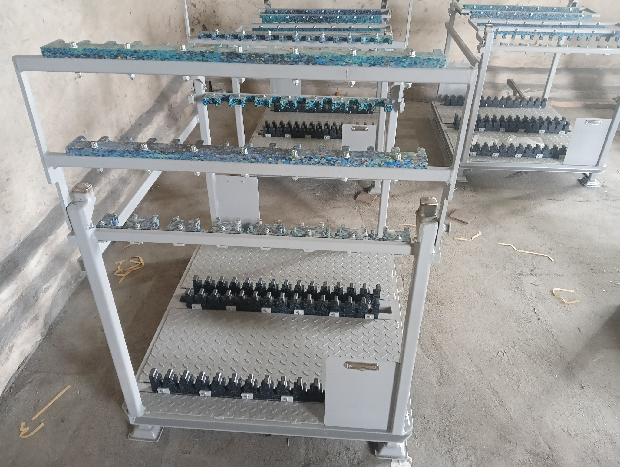 Metalowe wózki warsztatowe z półkami, na których umieszczone są detale z tworzywa sztucznego i metalu, prawdopodobnie przygotowane do obróbki.