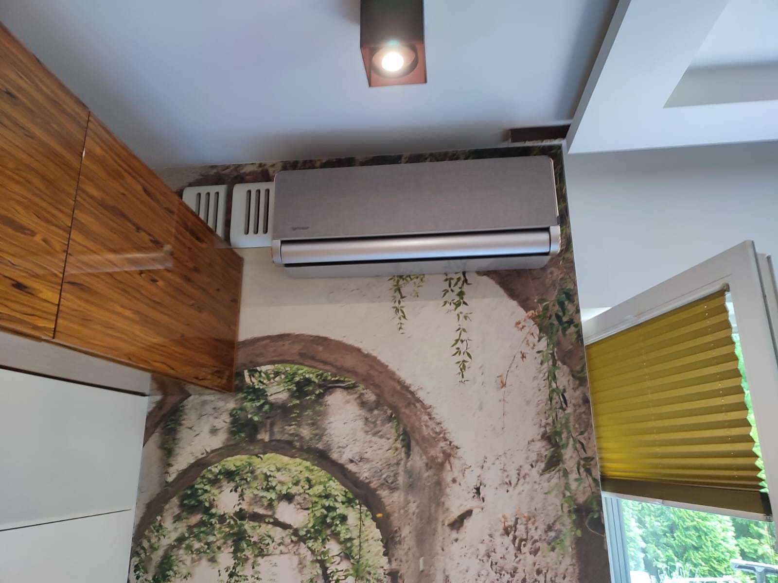 Szary klimatyzator ścienny zamontowany nad fototapetą z motywem ruin porośniętych bluszczem, widoczne fragmenty mebli kuchennych z połyskiem i okno z żaluzjami.