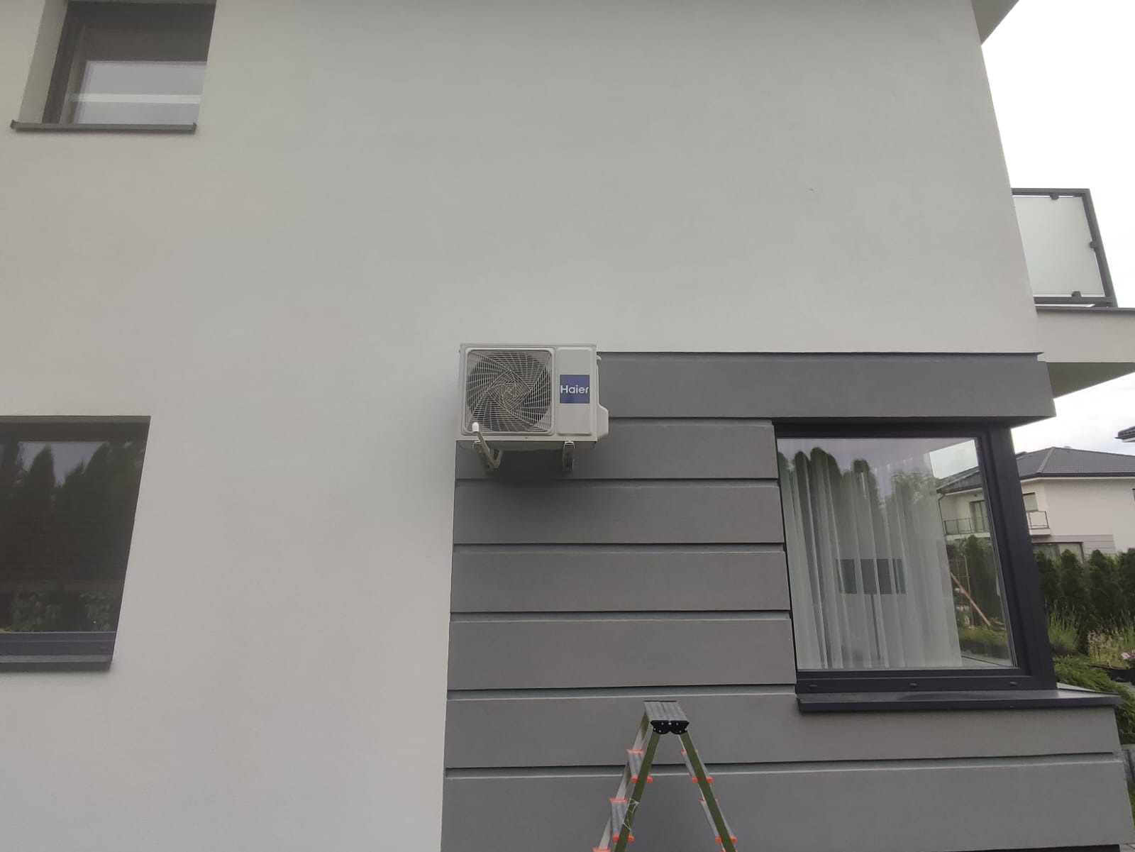 Zewnętrzna jednostka klimatyzacji marki Haier zamontowana na elewacji budynku, widoczna drabina oparta o ścianę.