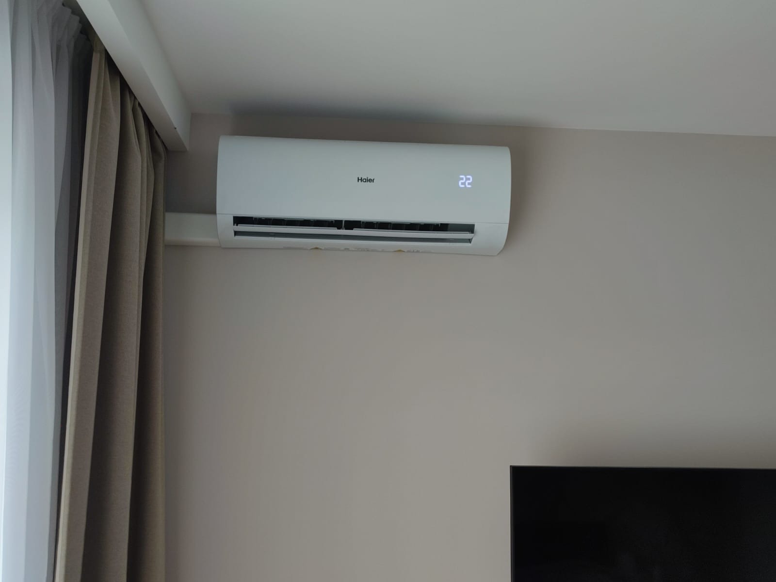 Biała jednostka klimatyzacyjna Haier zamontowana na ścianie w pokoju, wyświetlająca temperaturę 22 stopnie Celsjusza, z zasłoną w tle i fragmentem telewizora w dolnym rogu.