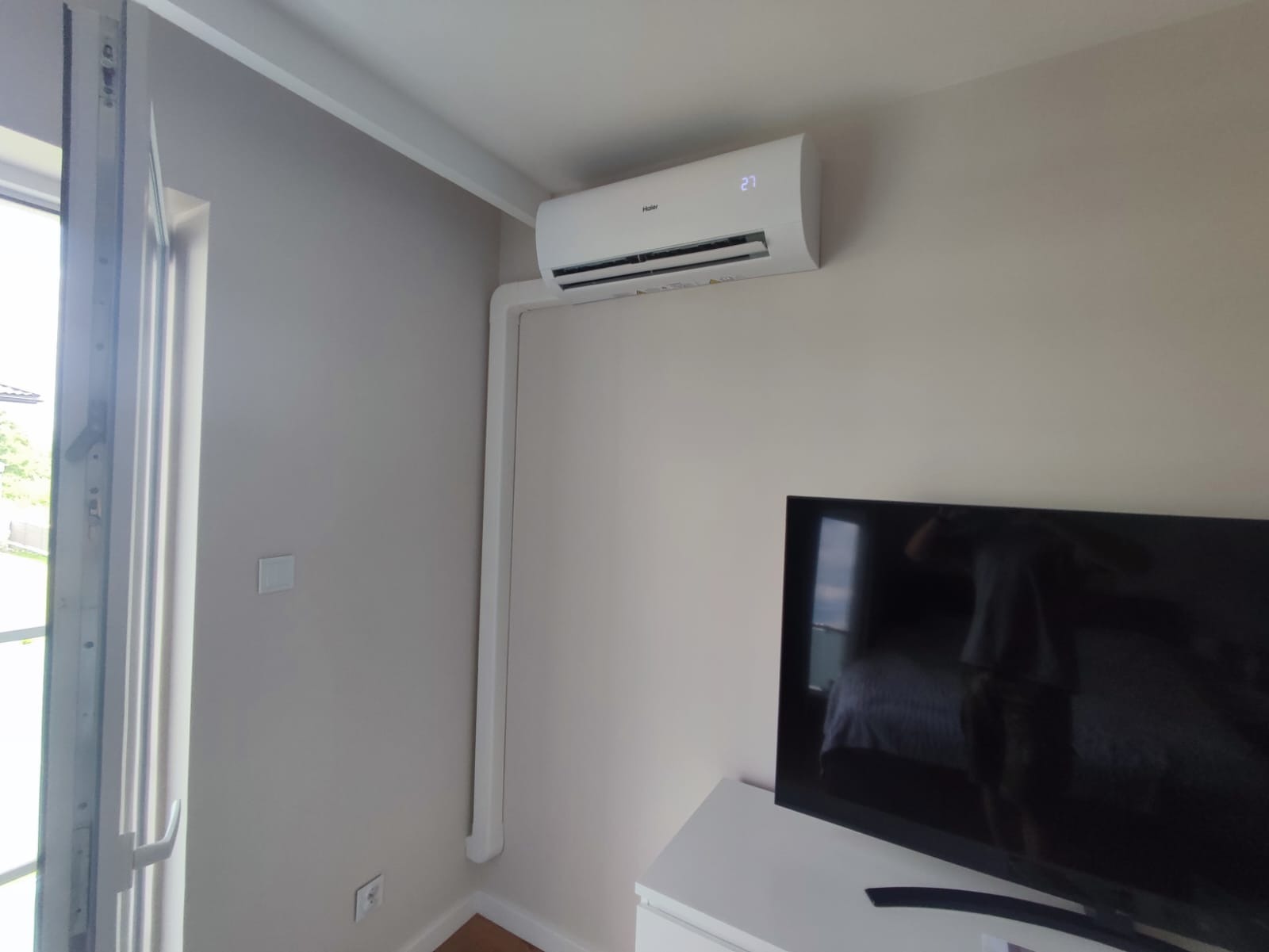 Biały klimatyzator marki Haier zamontowany na ścianie w pokoju, z widoczną białą listwą maskującą instalację, obok okna i telewizora.