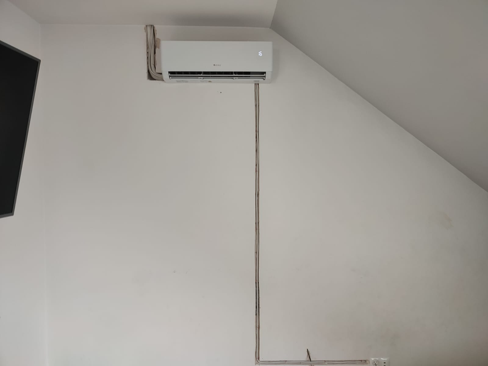 Biały klimatyzator zamontowany na skośnej ścianie poddasza z widocznymi przewodami w listwie przypodłogowej.