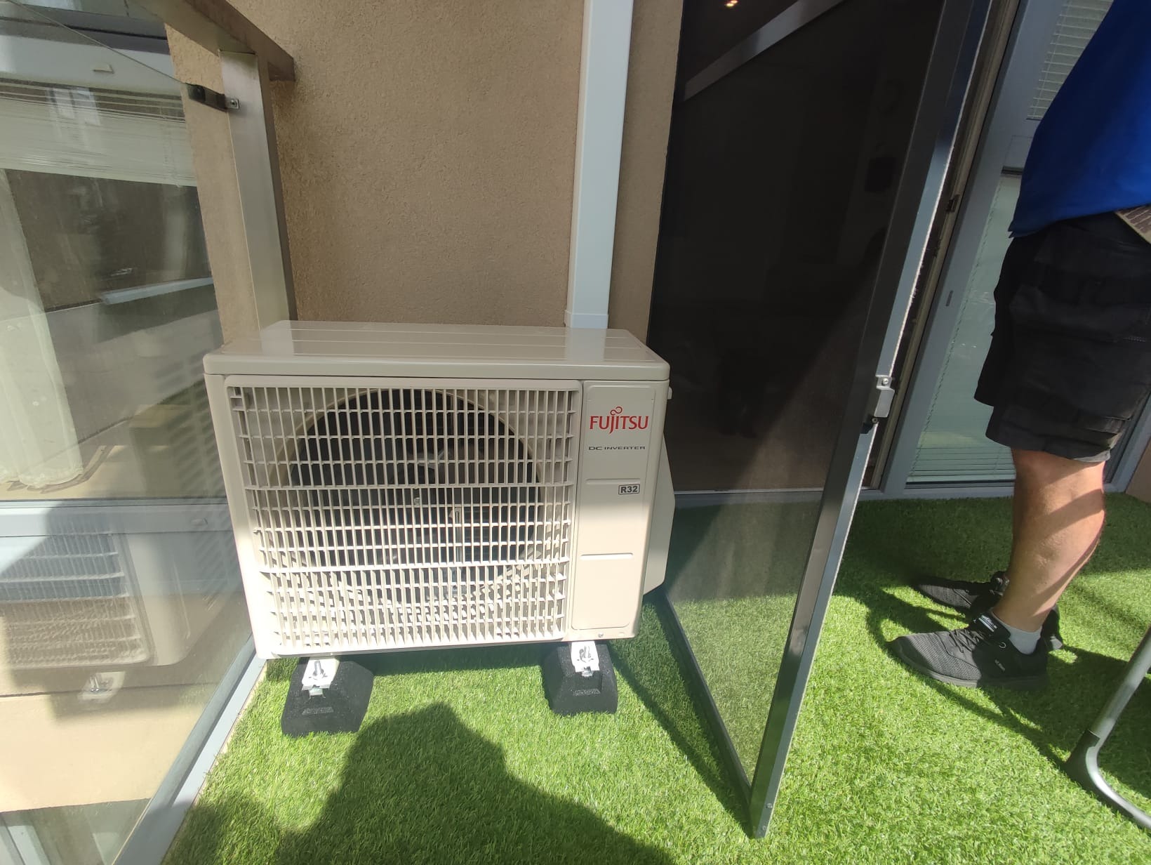 Zewnętrzna jednostka klimatyzacji Fujitsu DC Inverter R32 zamontowana na balkonie z zieloną sztuczną trawą, widoczna część osoby w tle.