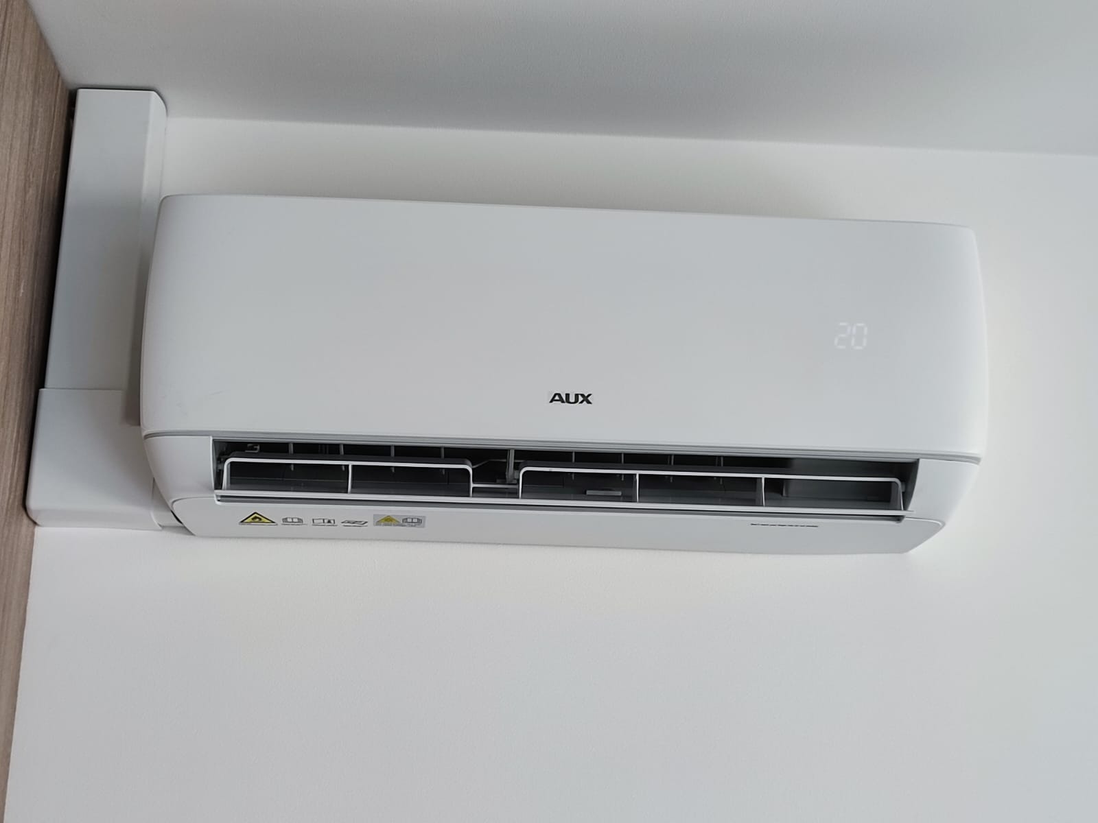 Biała klimatyzacja AUX zamontowana na białej ścianie, z widocznym wyświetlaczem temperatury ustawionym na 20 stopni Celsjusza.