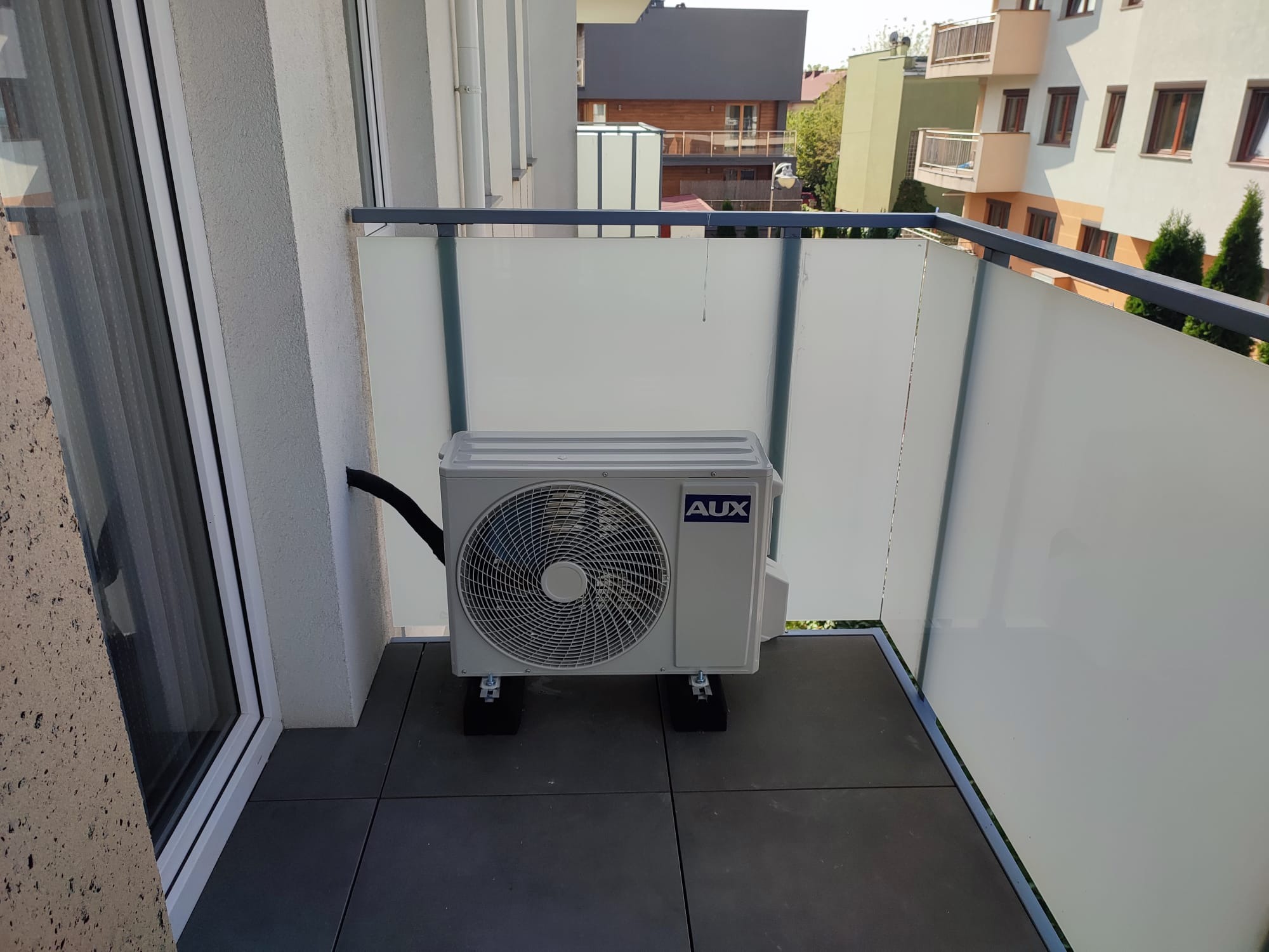 Jednostka zewnętrzna klimatyzacji AUX zamontowana na balkonie z szarymi płytkami i szklaną balustradą.