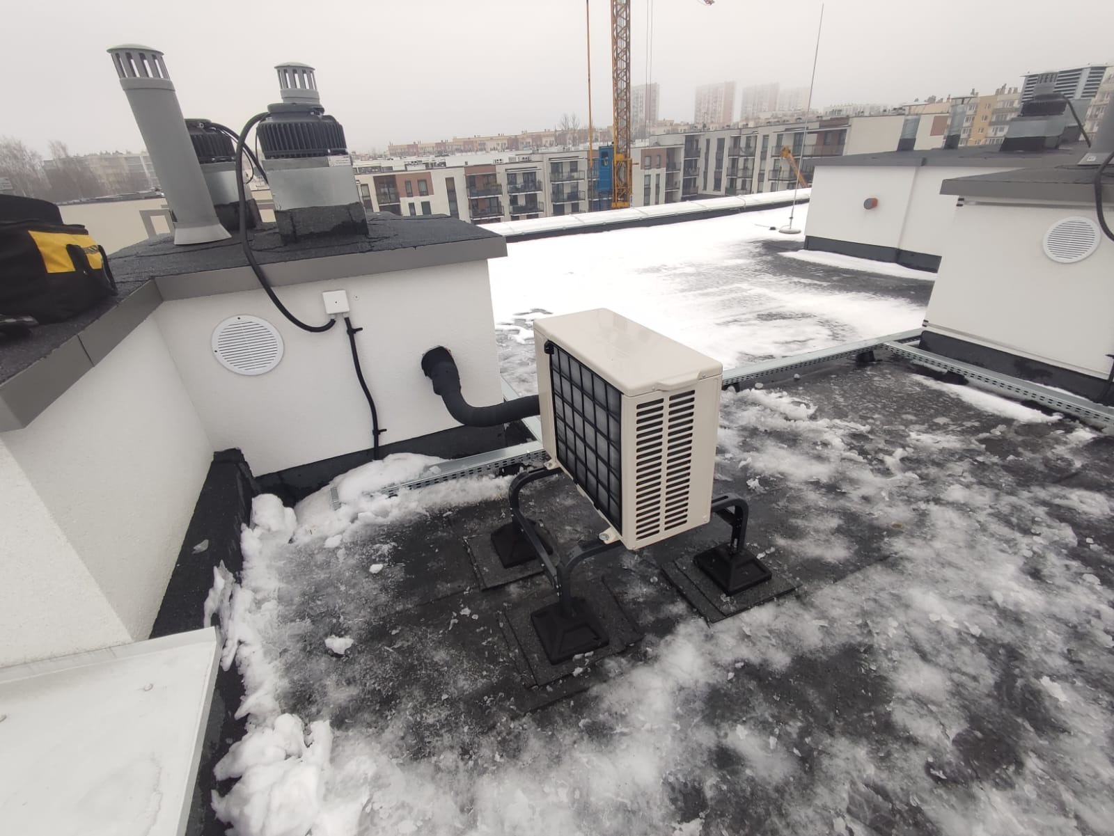 Klimatyzator zewnętrzny na płaskim dachu budynku wielorodzinnego, zamontowany na specjalnej konstrukcji, z widocznymi elementami instalacji i śniegiem na dachu.