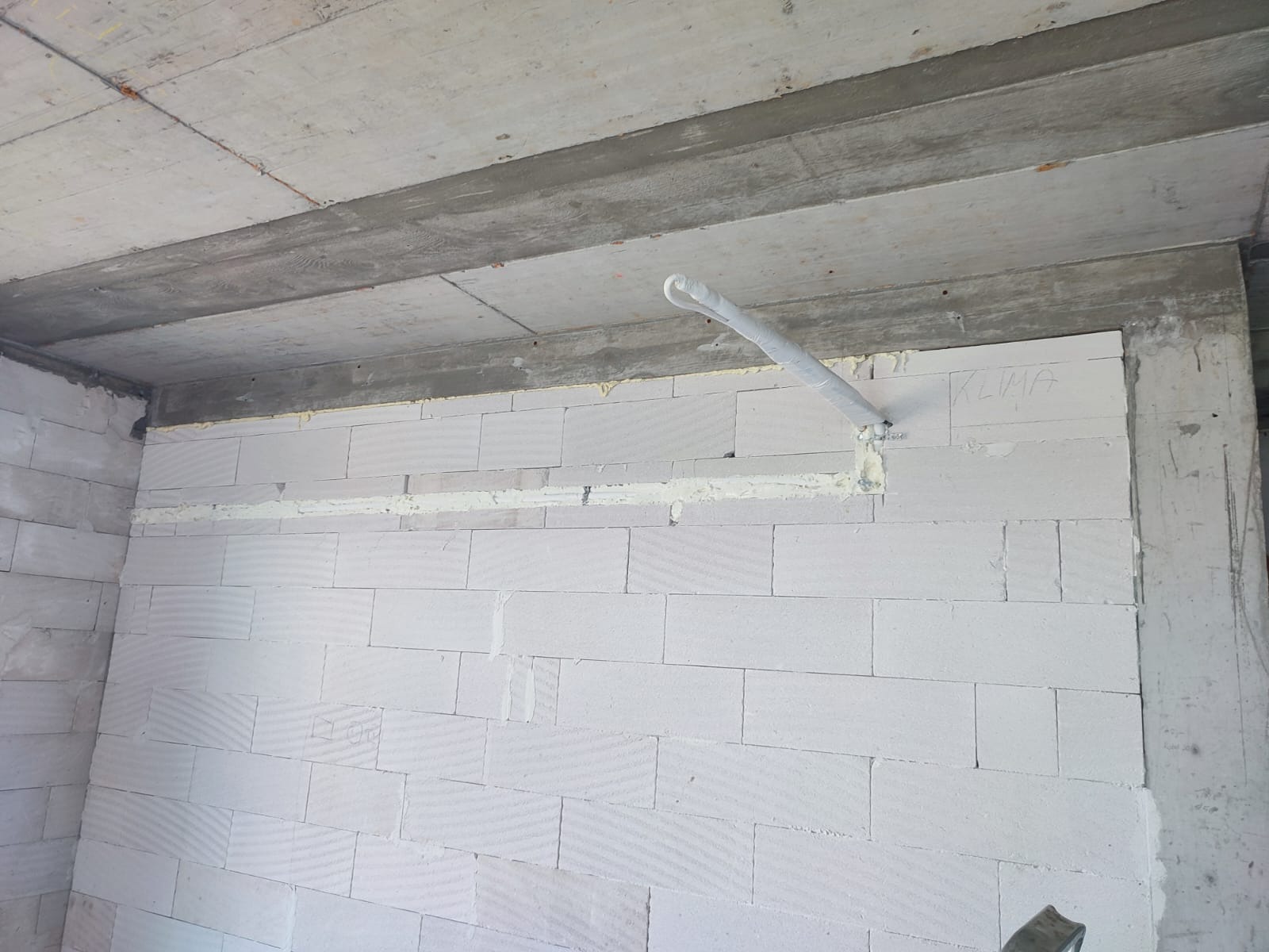 Ściana z betonu komórkowego z przygotowanym przepustem na rurę instalacji, zabezpieczoną białą folią i pianką montażową; napis 'KLIMA' naniesiony ołówkiem na ścianie.