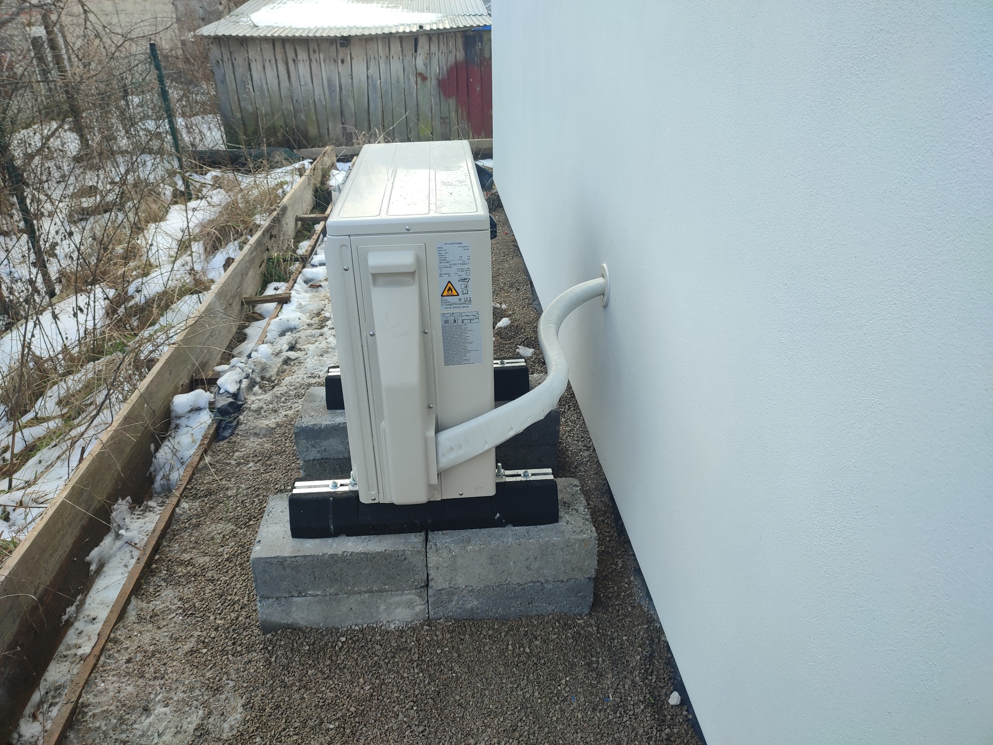 Zewnętrzna jednostka klimatyzatora zamontowana na betonowych bloczkach przy ścianie domu, z widoczną rurą odprowadzającą skropliny, na tle żwirowego podłoża i resztek śniegu.