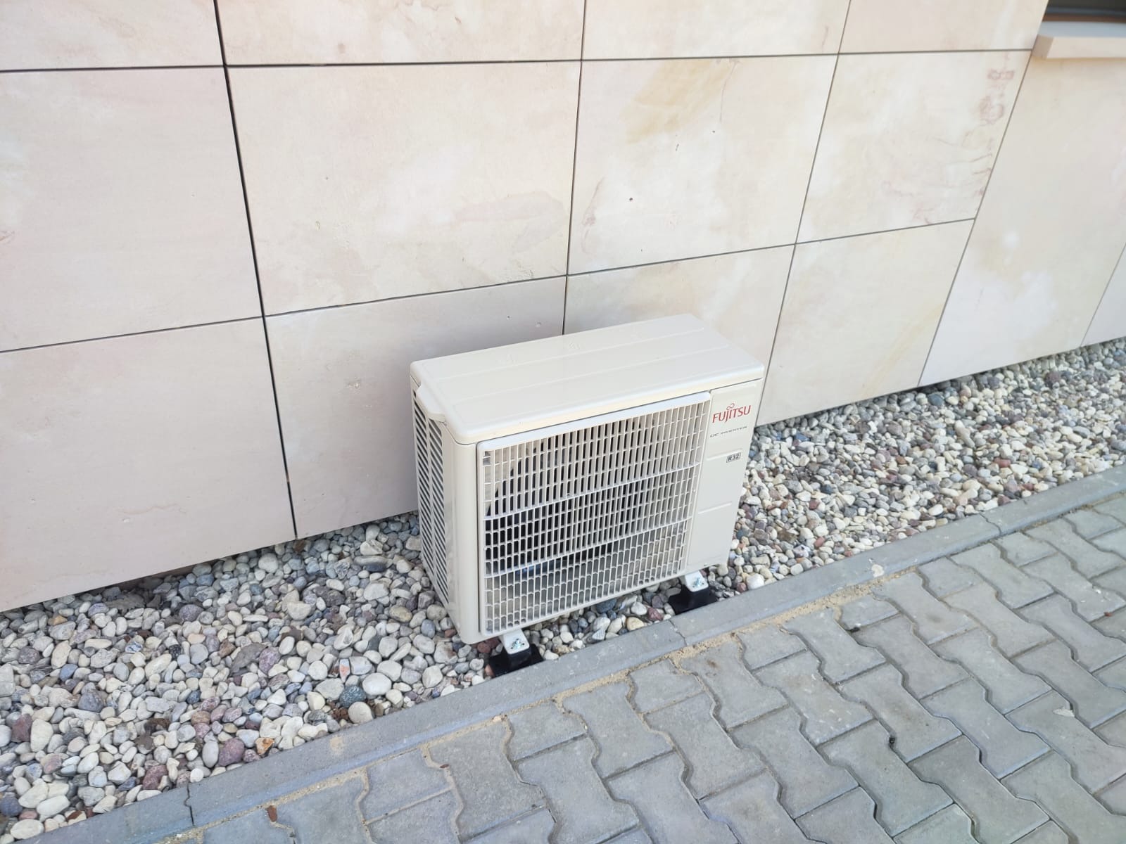 Zewnętrzna jednostka klimatyzacji Fujitsu zamontowana na elewacji wyłożonej jasnymi płytami, otoczona drobnymi kamieniami i kostką brukową.