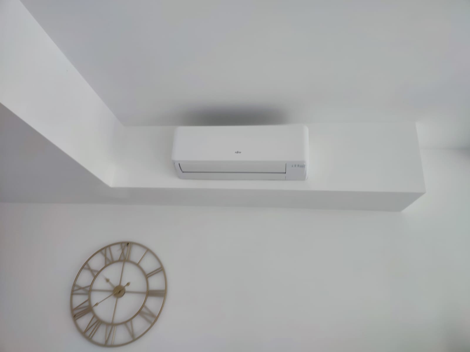 Biała klimatyzacja marki Fujitsu zamontowana na ścianie w nowoczesnym wnętrzu, obok dekoracyjny zegar z rzymskimi cyframi.