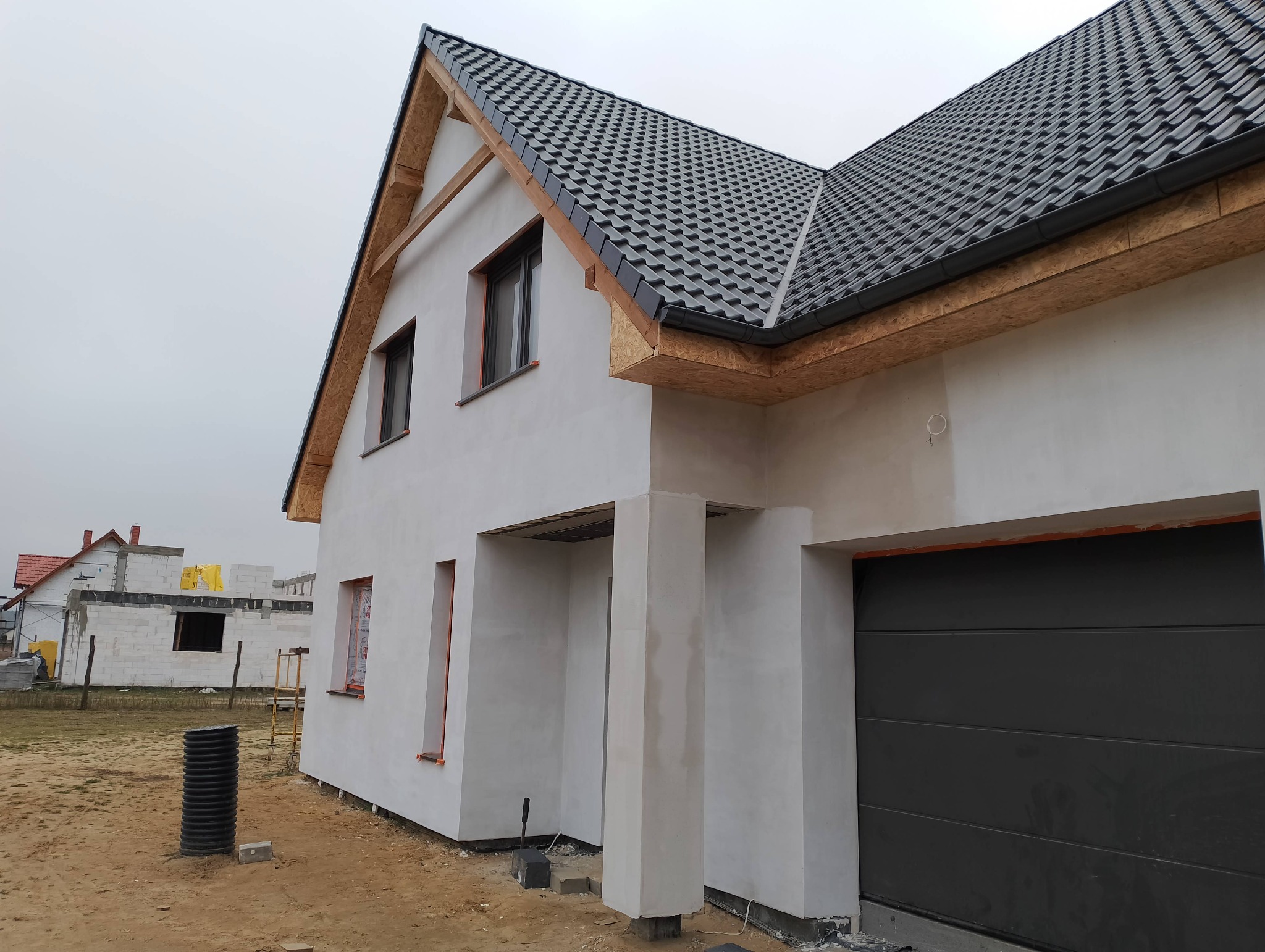 Surowy dom jednorodzinny w trakcie prac elewacyjnych, widoczne szare dachówki, biała elewacja w trakcie nakładania, okna z pomarańczowymi ramami, w tle budynek w trakcie budowy z żółtą folią ochronną.