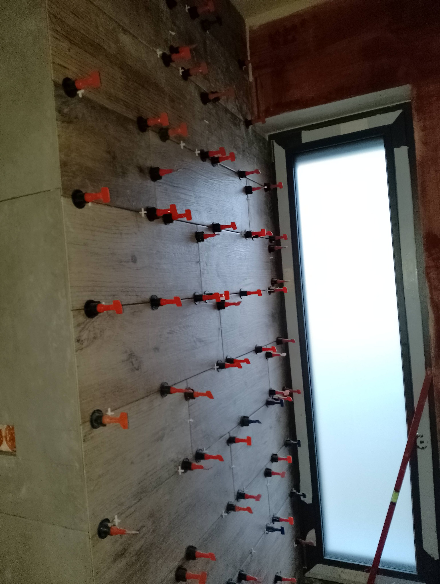 Ściana w trakcie układania płytek imitujących drewno, z użyciem systemu poziomowania płytek (klipsy i kliny), widoczny fragment okna i przygotowana ściana obok.