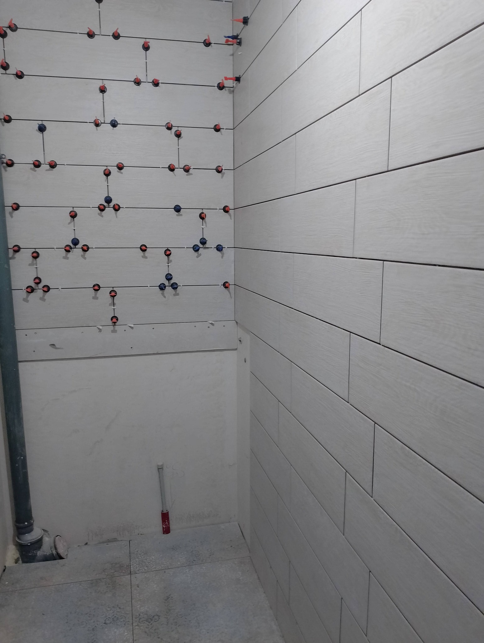 Ściana łazienki w trakcie remontu, wyłożona podłużnymi, szarymi płytkami imitującymi drewno, z widocznymi krzyżykami dystansowymi i systemem poziomowania płytek.