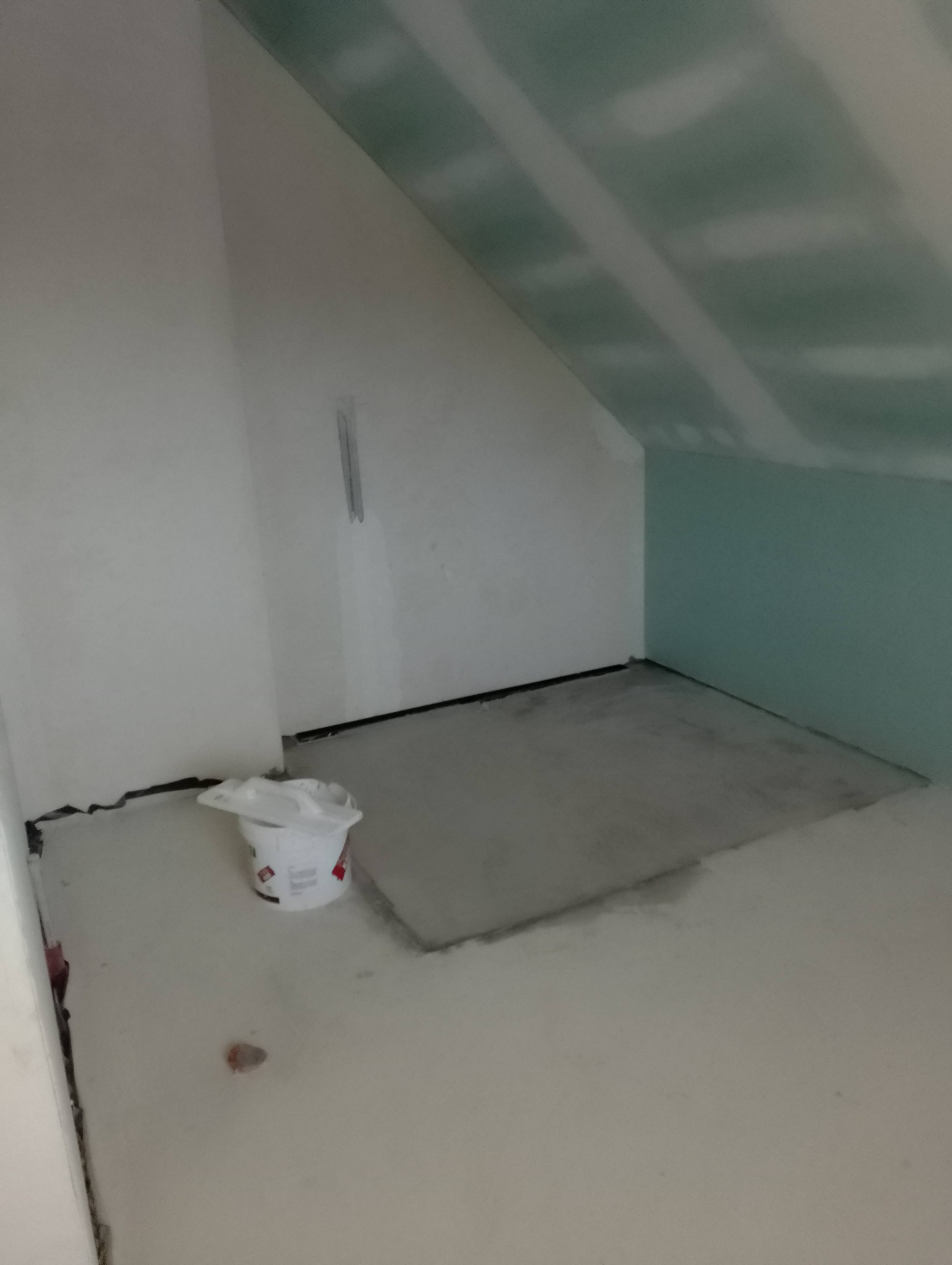 Poddasze w trakcie remontu, widoczne płyty gipsowo-kartonowe na skosach, szpachlowane ściany, wiadro farby i fragment wylewki na podłodze.