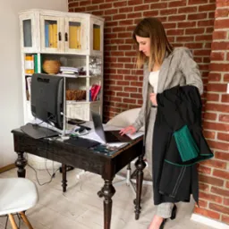 Kobieta w garniturze w kratę stoi przy biurku z laptopem, trzymając togę z zielonym żabotem na tle ściany z czerwonej cegły.