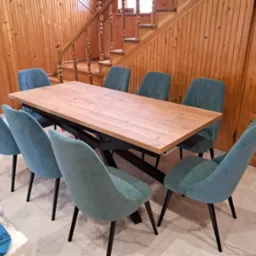 Drewniany stół z czarną podstawą i ośmioma turkusowymi krzesłami na tle drewnianej ściany i schodów. Nowoczesny design z naturalnymi materiałami.