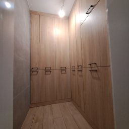 F.H.U Kamix Kamil Zieliński - Wnętrze garderoby z jasnymi, drewnianymi szafami wnękowymi o minimalistycznych, czarnych uchwytach. Widok z perspektywy wejścia, oświetlenie sufitowe.