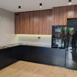 Nowoczesna kuchnia z drewnianymi szafkami górnymi i czarnymi dolnymi, blatem z ciemnego kamienia i wbudowanym sprzętem AGD. Widoczne refleksy w lodówce.