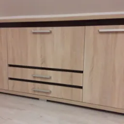 Jasna komoda z szufladami i szafkami, wykończona w okleinie imitującej drewno, z prostymi, metalowymi uchwytami, ustawiona na jasnej podłodze.