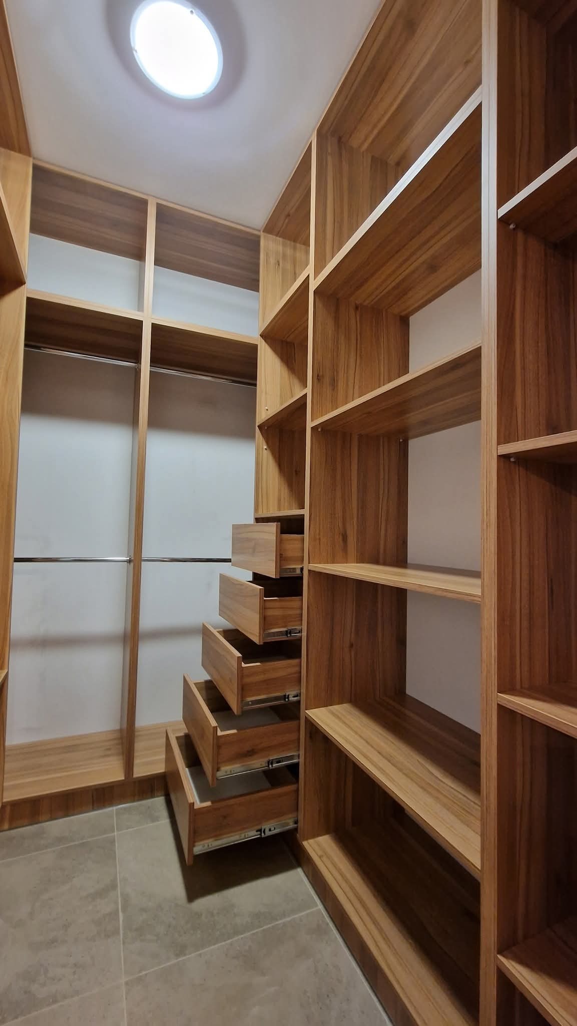 Drewniana szafa wnękowa z otwartymi półkami i wysuwanymi szufladami, oświetlona od góry, na tle jasnej ściany i podłogi z płytek.