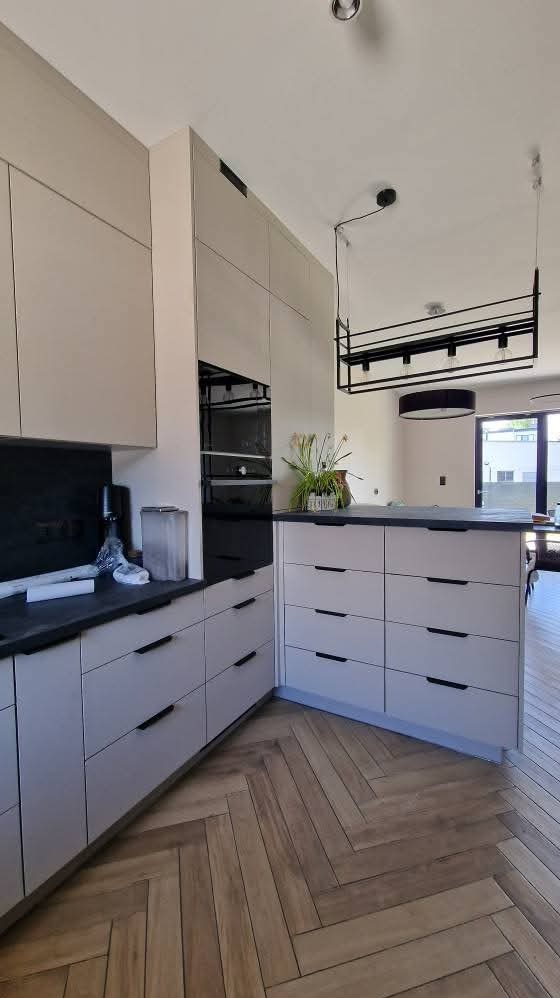 Jasna kuchnia z drewnianą podłogą w jodełkę. Zabudowa kuchenna z szufladami i piekarnikiem. Nad wyspą kuchenną wisi nowoczesna lampa.