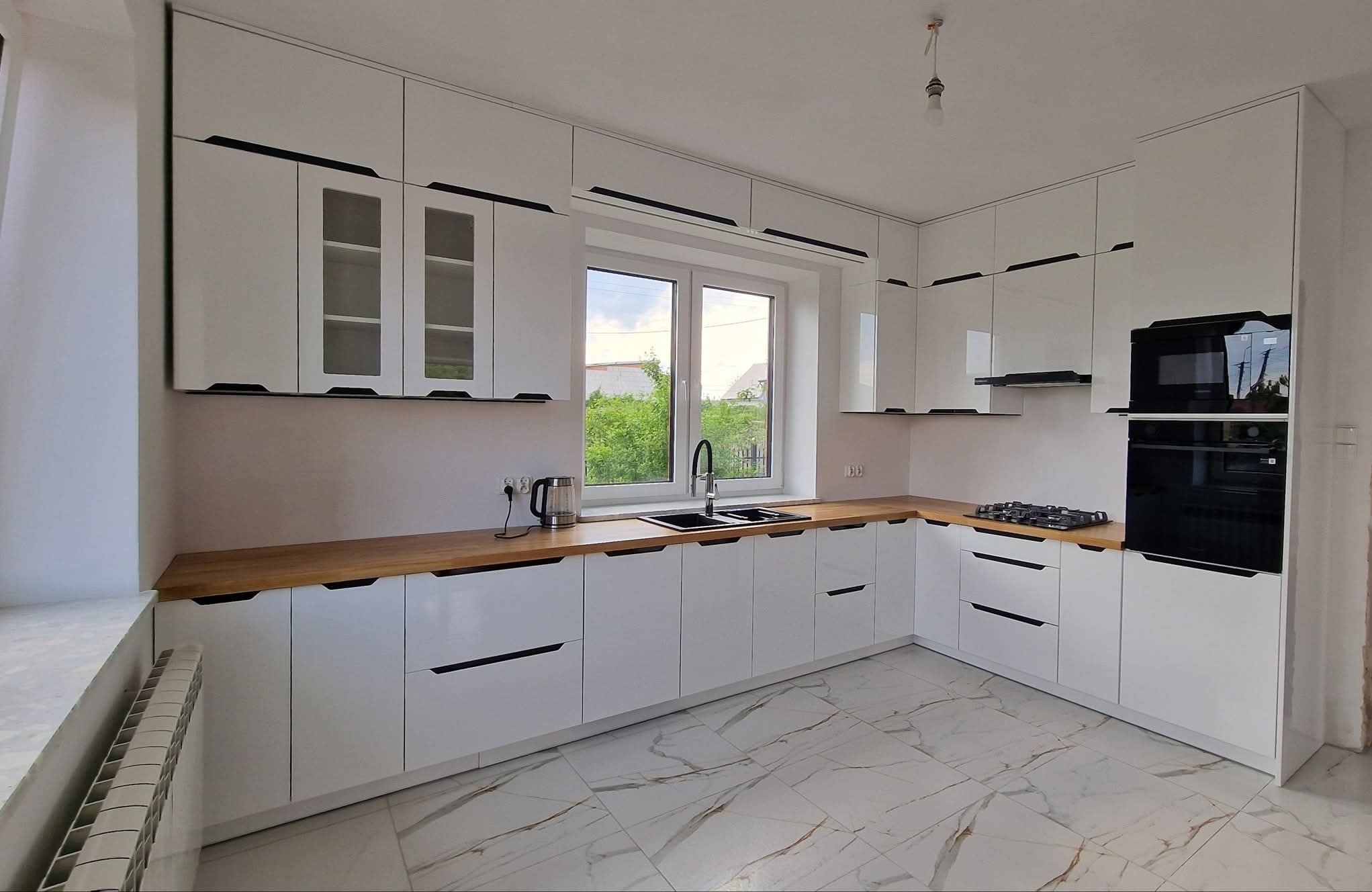 Nowoczesna kuchnia z białymi lakierowanymi frontami, drewnianym blatem i czarnymi uchwytami, widok na okno i marmurowe płytki podłogowe.