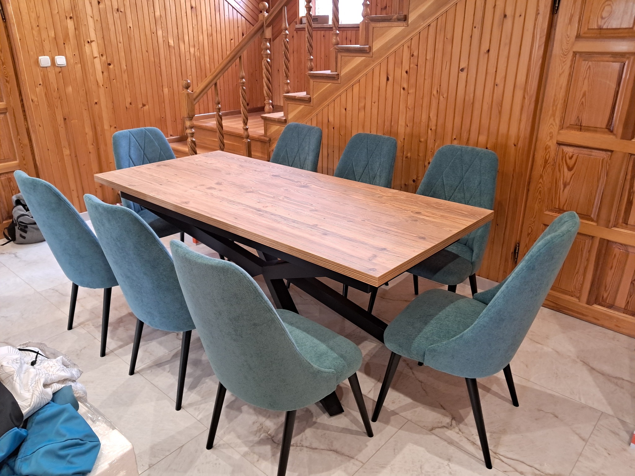 Drewniany stół z czarną podstawą i ośmioma turkusowymi krzesłami na tle drewnianej ściany i schodów. Nowoczesny design z naturalnymi materiałami.