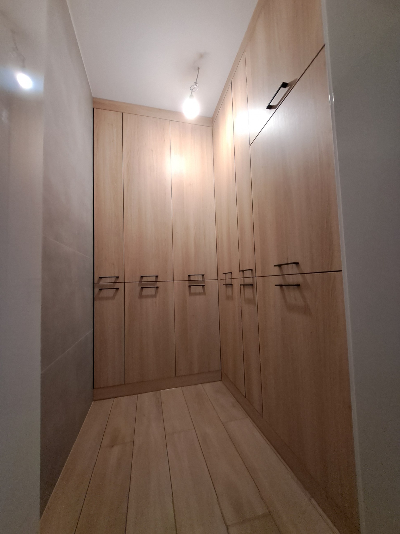 Wnętrze garderoby z jasnymi, drewnianymi szafami wnękowymi o minimalistycznych, czarnych uchwytach. Widok z perspektywy wejścia, oświetlenie sufitowe.