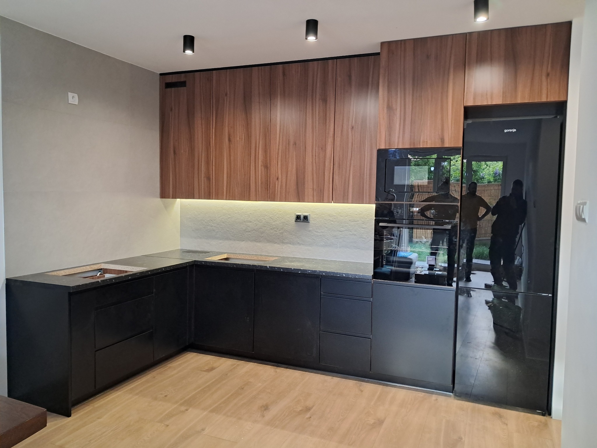 Nowoczesna kuchnia z drewnianymi szafkami górnymi i czarnymi dolnymi, blatem z ciemnego kamienia i wbudowanym sprzętem AGD. Widoczne refleksy w lodówce.