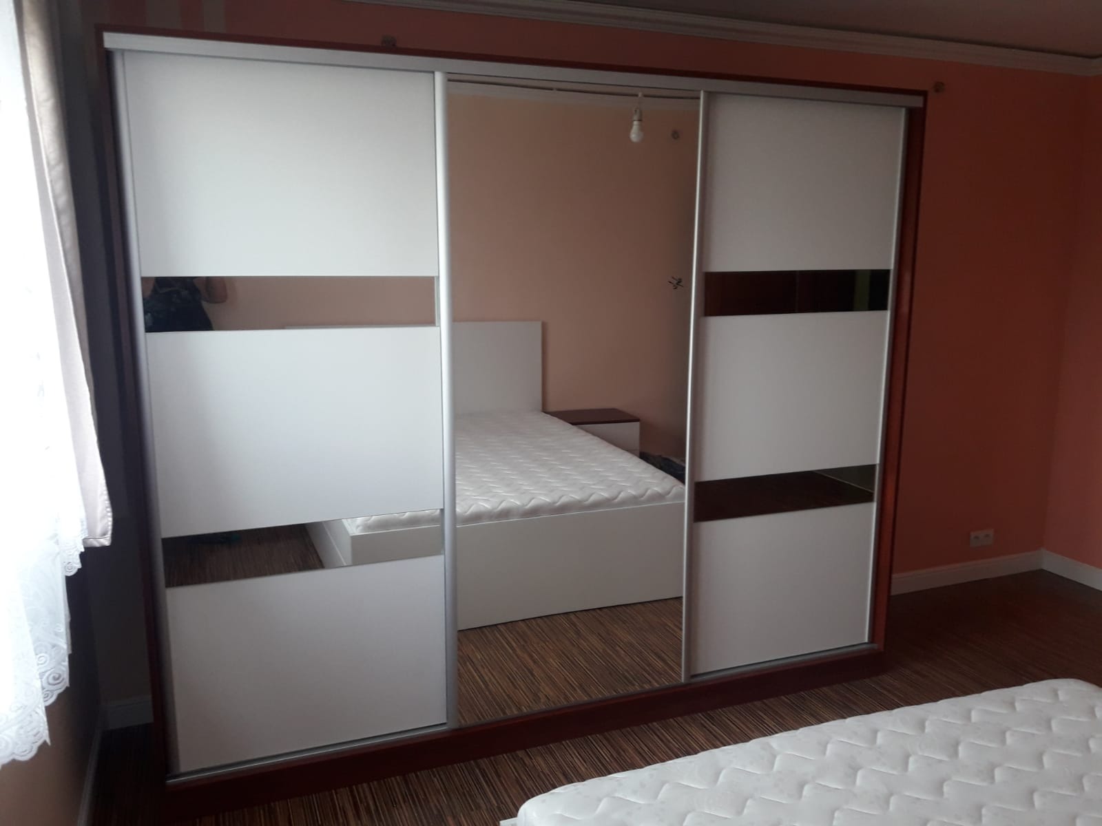 Trzydrzwiowa szafa przesuwna z białymi frontami i poziomymi pasami lustrzanymi, odbijająca wnętrze sypialni z łóżkiem i szafką nocną.