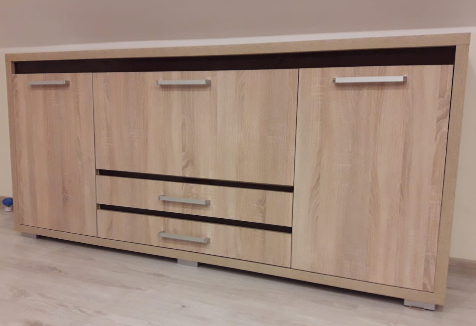 Jasna komoda z szufladami i szafkami, wykończona w okleinie imitującej drewno, z prostymi, metalowymi uchwytami, ustawiona na jasnej podłodze.