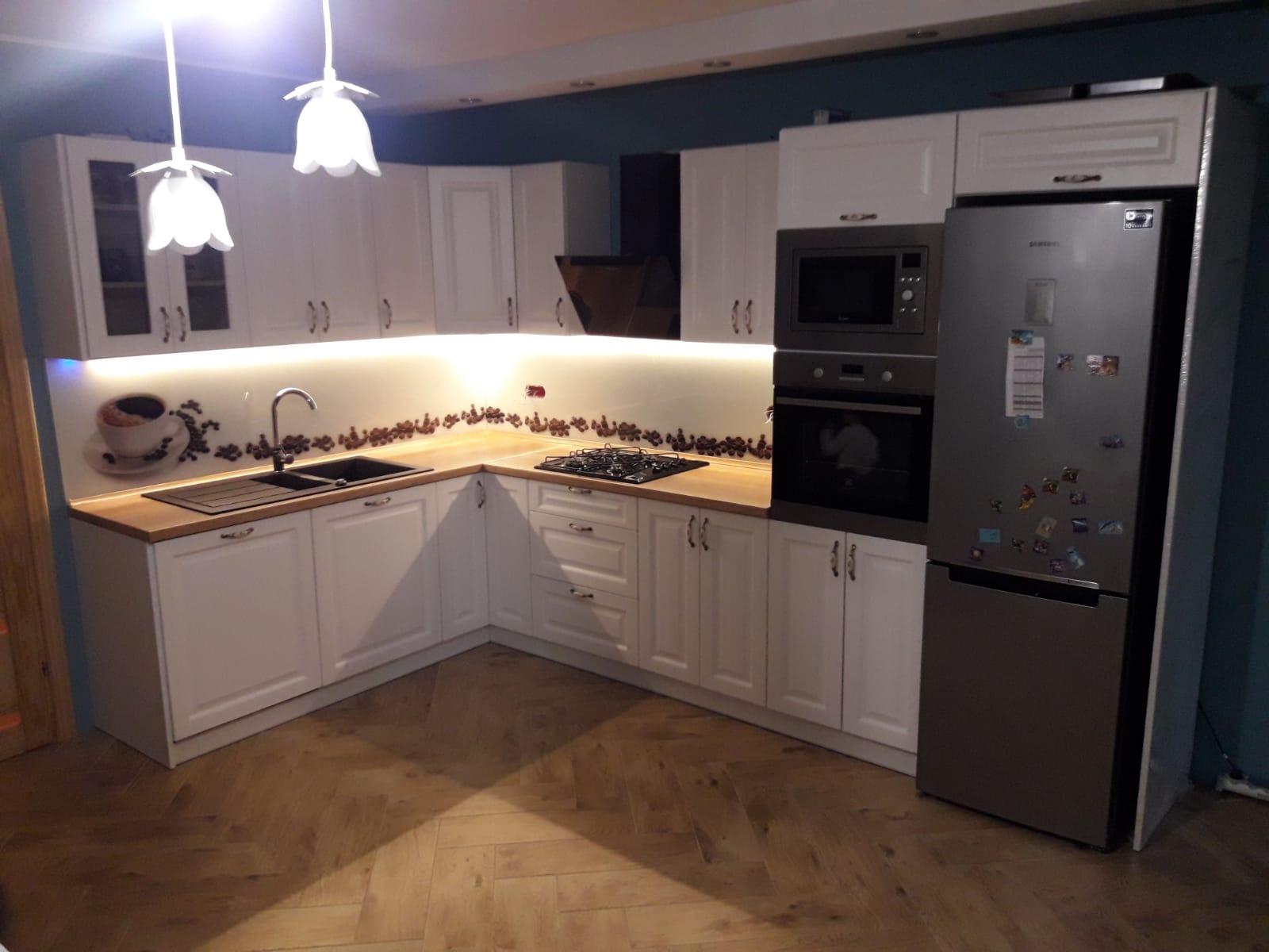 Jasna, narożna kuchnia z białymi szafkami, drewnianym blatem, płytą grzewczą i zlewozmywakiem, oświetlona lampami w kształcie kwiatów, z motywem kawy na ścianie i parkietem w jodełkę.