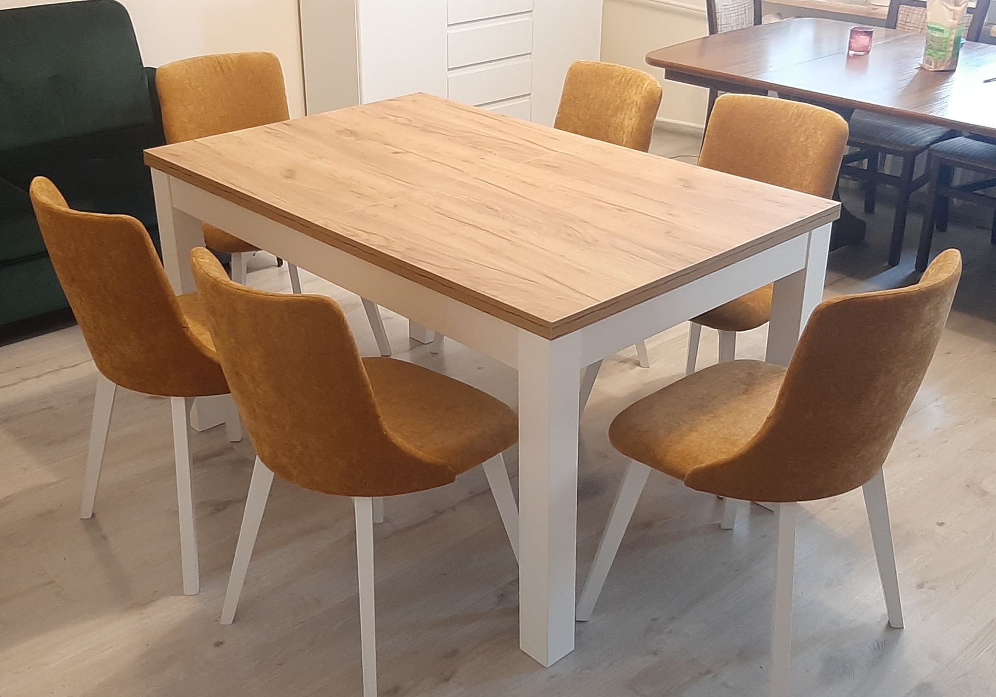 Stół z blatem imitującym jasne drewno, wsparty na białych nogach, otoczony sześcioma krzesłami z tapicerką w odcieniach musztardowej żółci, na tle fragmentu zielonej sofy i białej komody.