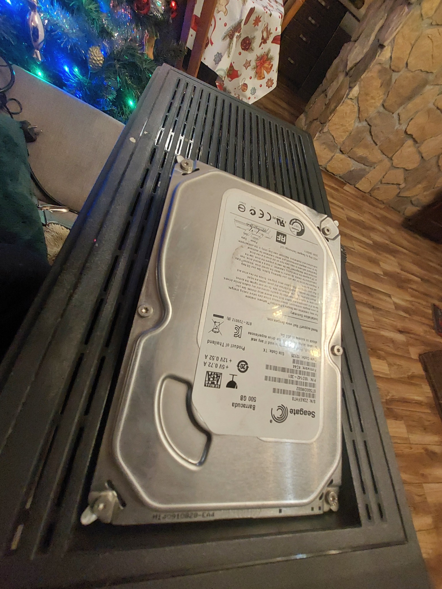 500 GB Seagate Barracuda dysk twardy, leżący na czarnej, perforowanej obudowie komputera, w tle widoczna choinka i kamienna ściana.