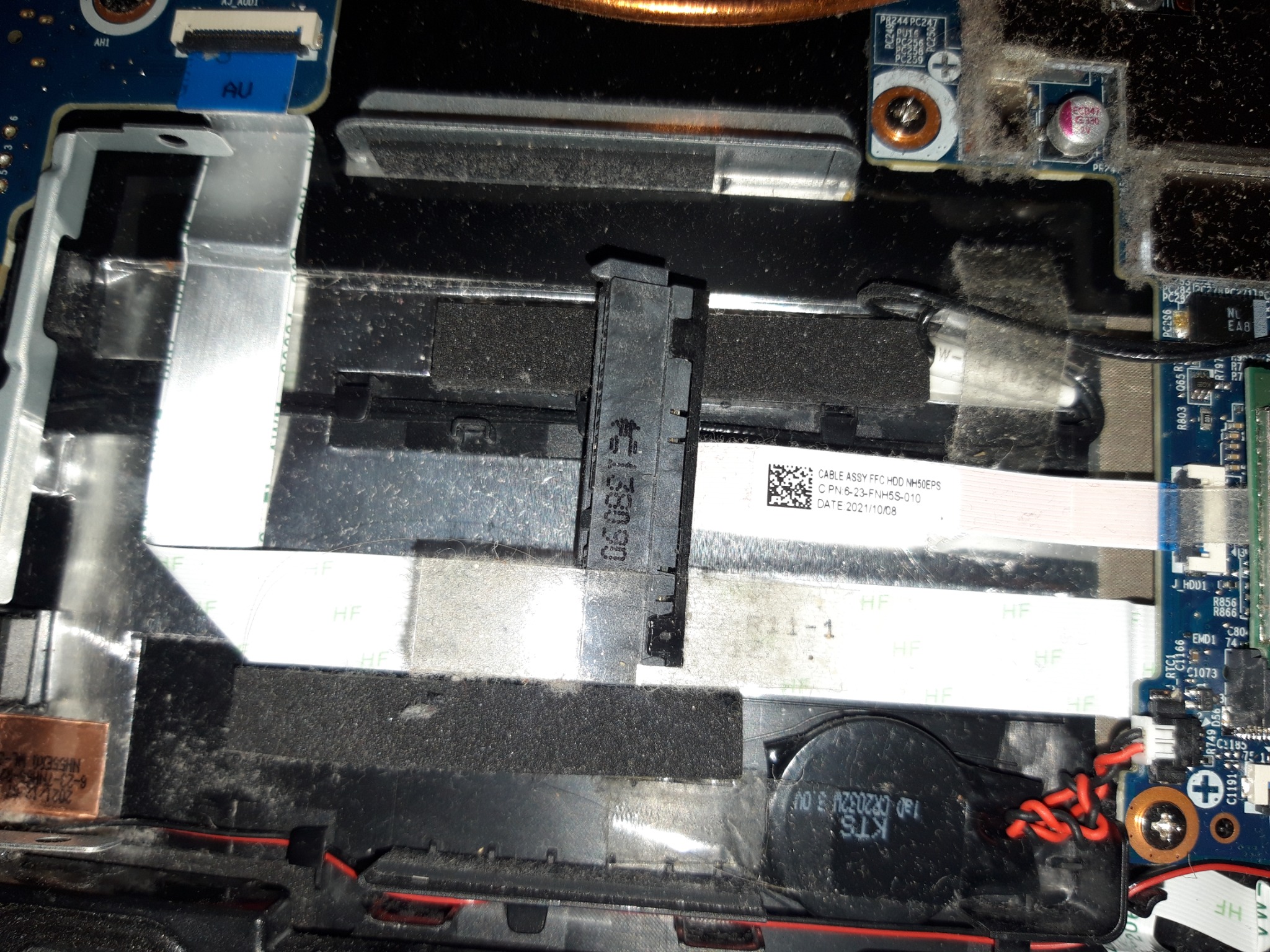 Wnętrze brudnego laptopa z widocznymi elementami płyty głównej, kablami taśmowymi z oznaczeniami HF, małą baterią CR2032 i fragmentem wentylatora, ukazujące stan przed czyszczeniem lub naprawą.