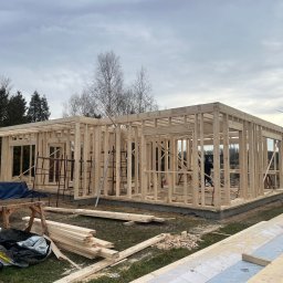 KUBIKDOM - Konstrukcja domu w technologii szkieletowej w trakcie budowy. Widoczne drewniane elementy konstrukcyjne, teren budowy z materiałami i drabina. Pochmurne niebo w tle.