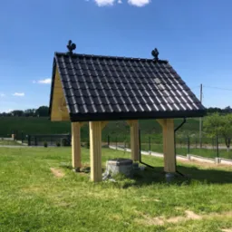 Zadaszenie studni z czarną dach&oacute;wką i ozdobnymi kogutami na szczycie, wsparte na jasnych, drewnianych słupach, otoczone zielonym trawnikiem i elementami instalacji wodnej.