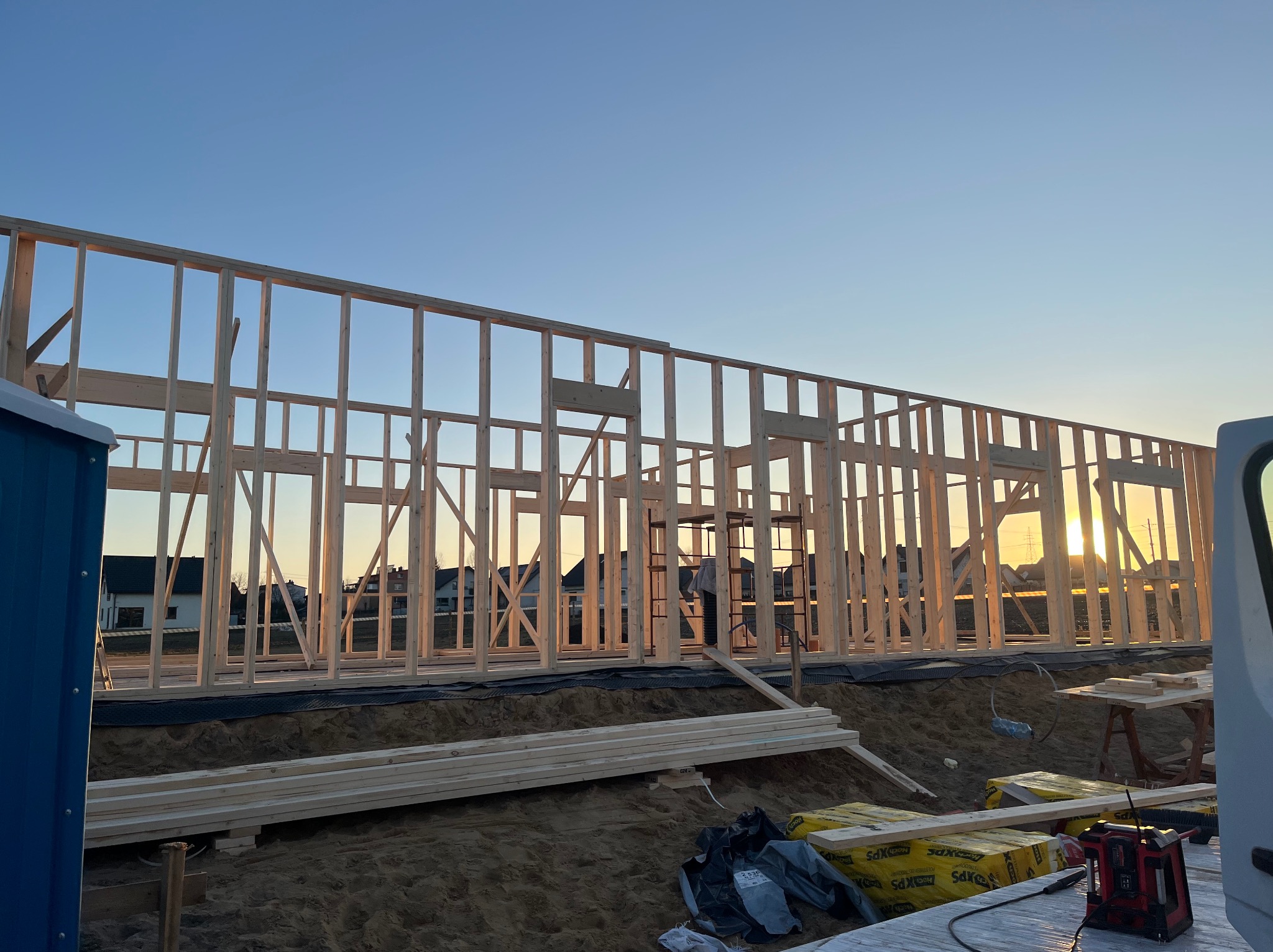 Konstrukcja szkieletowa domu w trakcie budowy o zachodzie słońca. Widoczne drewniane elementy i materiały budowlane. Częściowo widoczny przenośny WC.