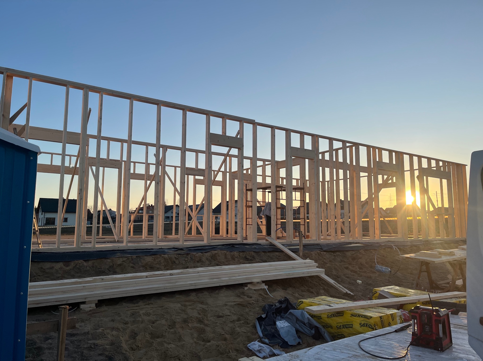 Konstrukcja drewniana domu w trakcie budowy, widok na szkielet budynku na tle zachodzącego słońca. Na pierwszym planie materiały budowlane i narzędzia.