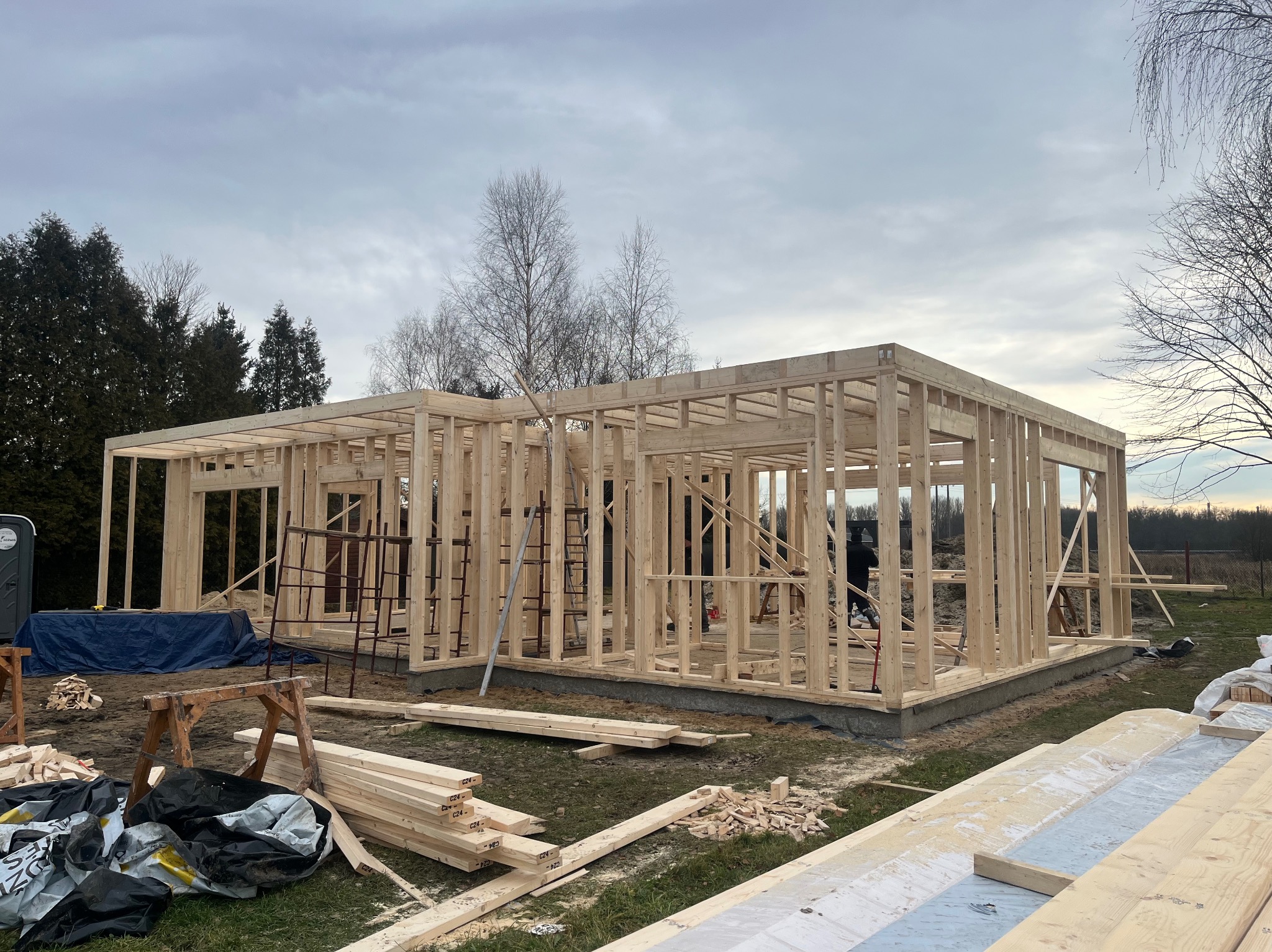 Konstrukcja domu w technologii szkieletowej w trakcie budowy. Widoczne drewniane elementy konstrukcyjne, teren budowy z materiałami i drabina. Pochmurne niebo w tle.