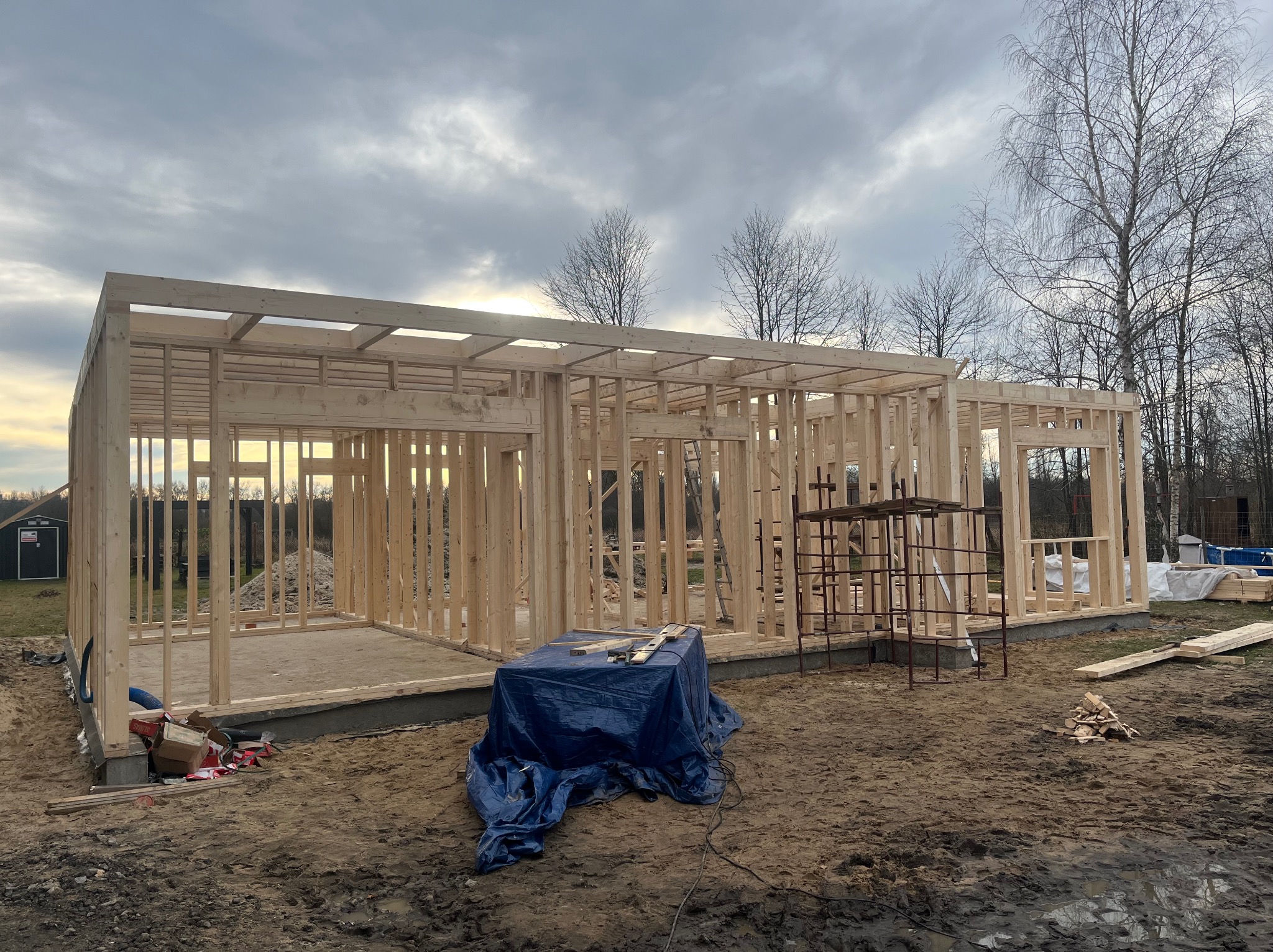 Surowy szkielet domu w budowie, drewniane belki tworzą ściany i zarys dachu na tle pochmurnego nieba. Widoczne narzędzia i materiały budowlane.