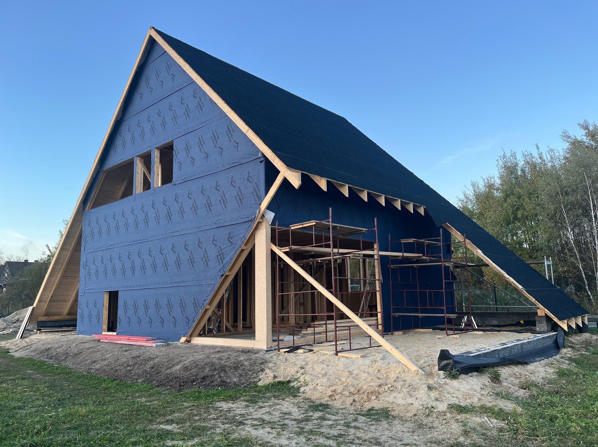 Wznoszenie domu szkieletowego w kształcie litery A. Widoczne rusztowania, folia ochronna i fragment dachu. Konstrukcja w otoczeniu zieleni i piasku.