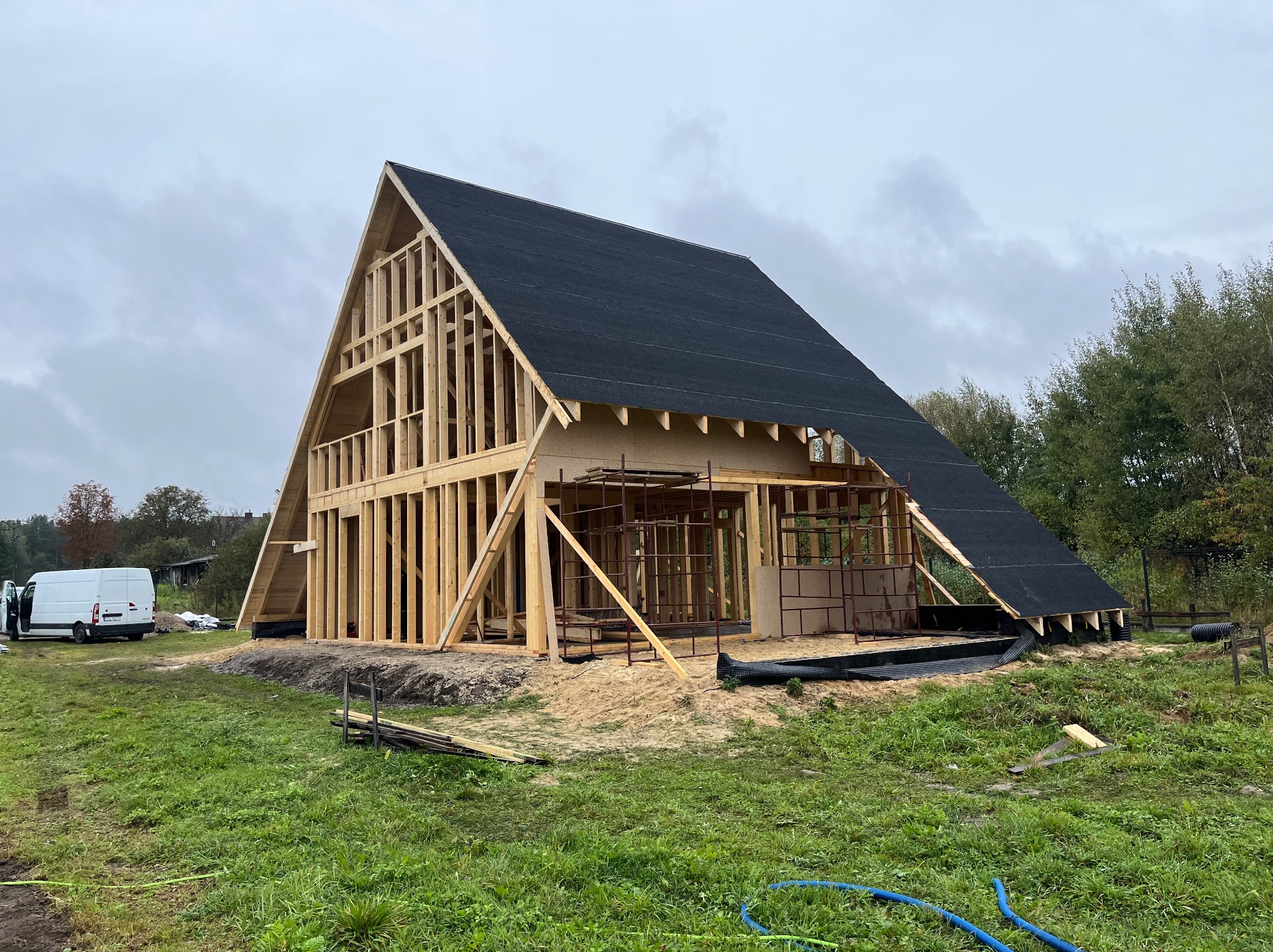 Wznoszenie domu szkieletowego z dachem pokrytym papą. Widoczna drewniana konstrukcja, rusztowania i elementy izolacyjne. Na trawie leżą węże. W tle biały bus.