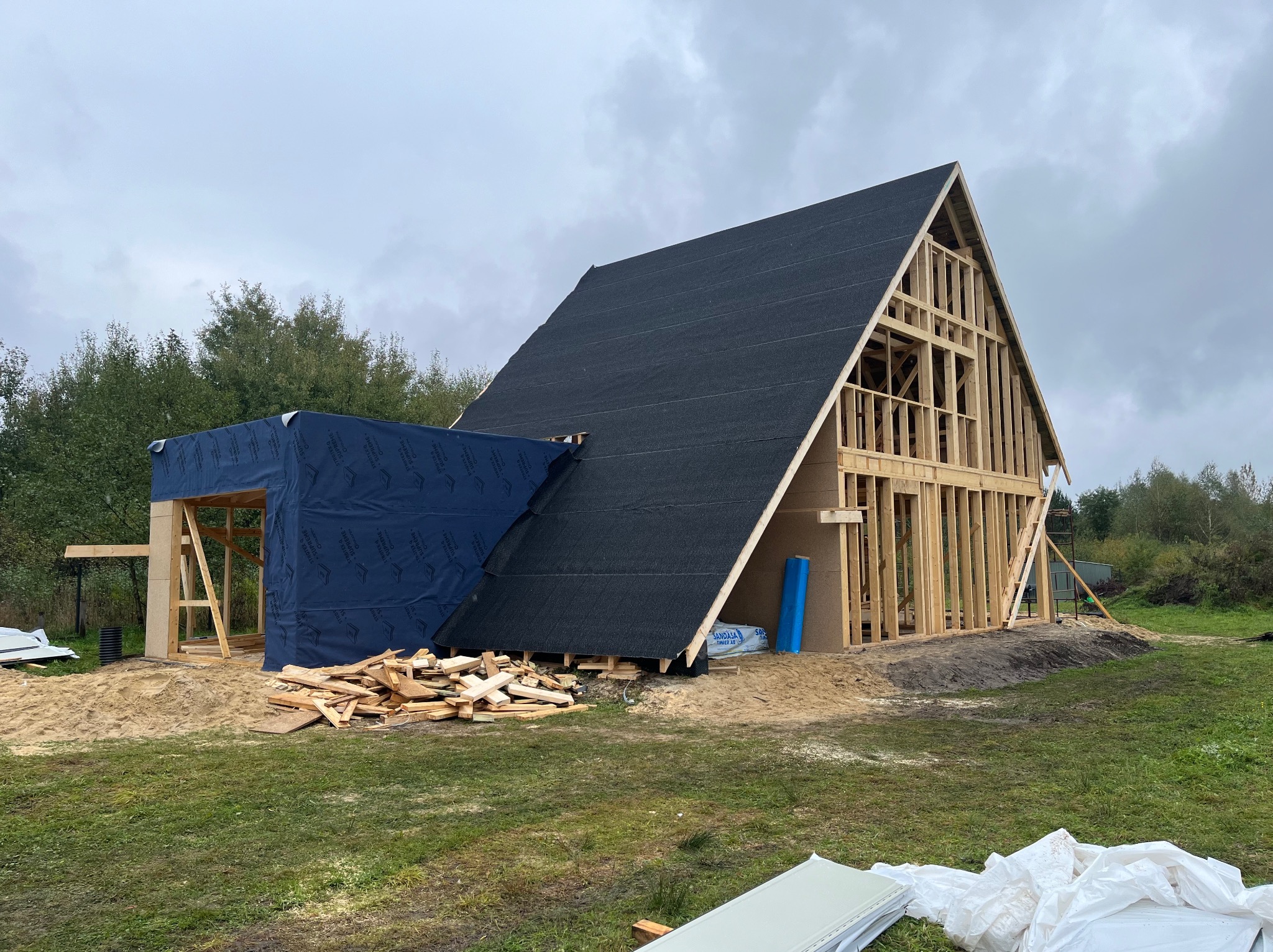 Budowa domu szkieletowego w trakcie: widoczna drewniana konstrukcja, czarny dach pokryty papą, niebieska membrana i stos drewna. Etap wznoszenia domu.