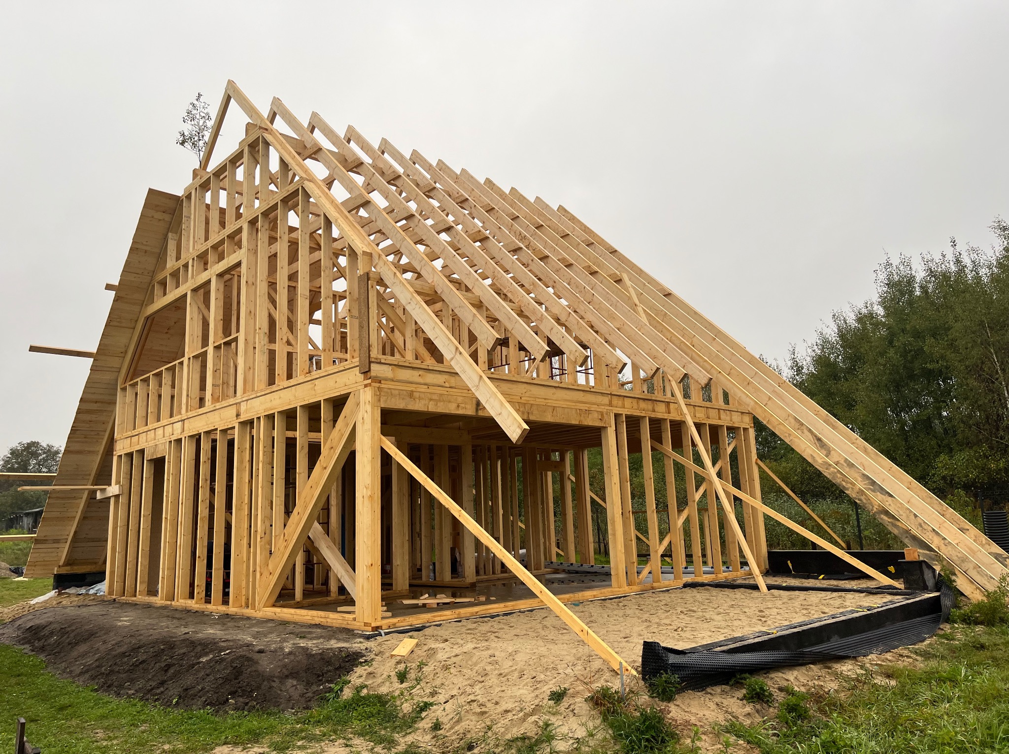 Surowa konstrukcja domu w systemie szkieletowym, widoczna drewniana rama i krokwie dachowe na tle pochmurnego nieba i drzew, w trakcie budowy.