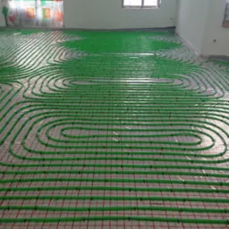 Zielone rury ogrzewania podłogowego ułożone na siatce z czerwonymi liniami w jasnym pomieszczeniu w trakcie montażu.