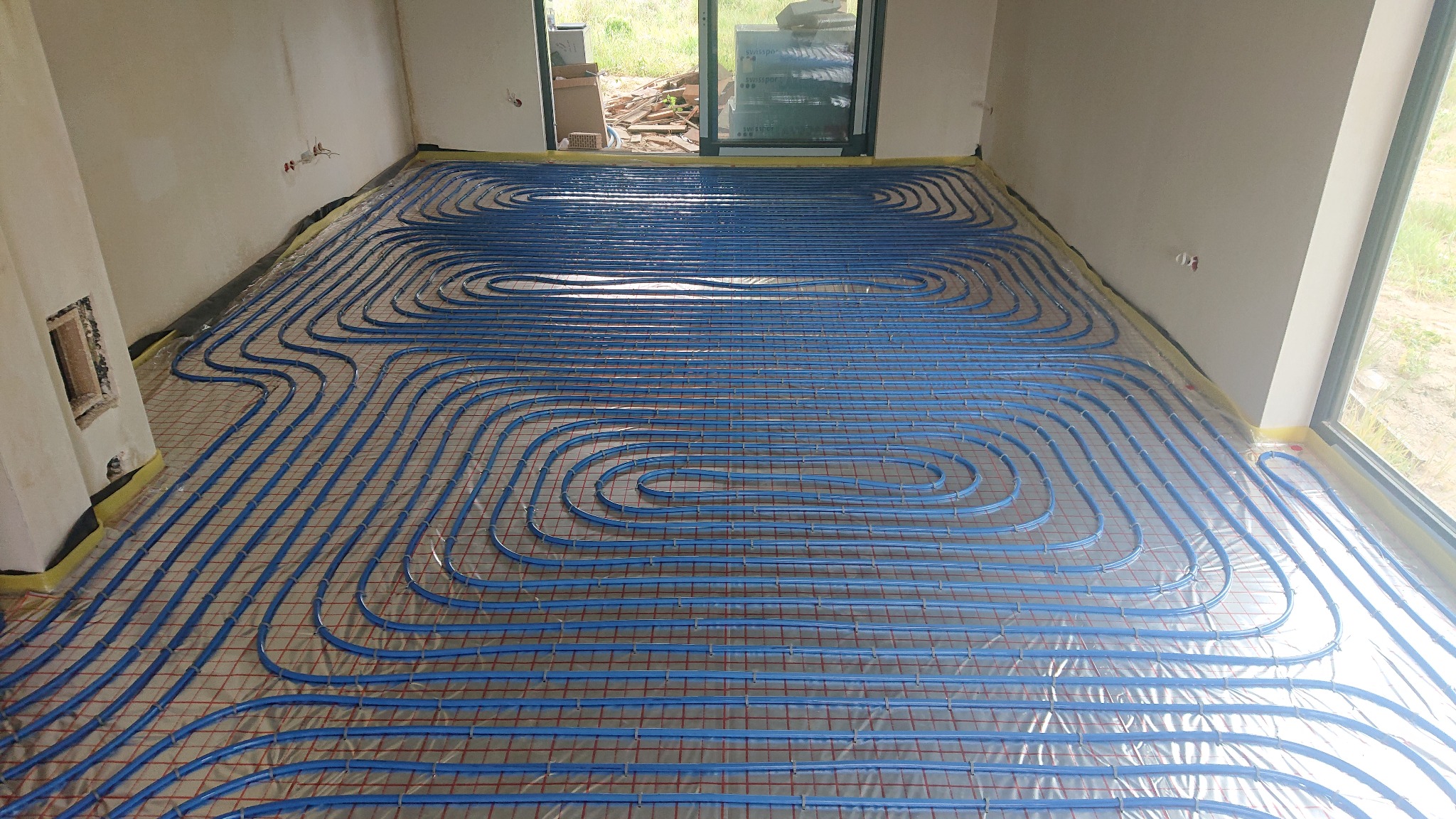 Niebieskie rury ogrzewania podłogowego ułożone na czerwonej siatce w surowym wnętrzu z widokiem na zewnątrz przez duże okno.